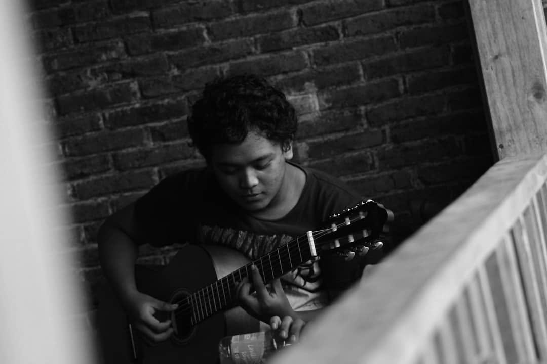 Black and white photo of a young man playing an acoustic guitar by a brick wall